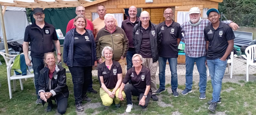 Dreizehn Boule-Spieler und Spielerinnen in Vereinskleidung vor Holzhütte