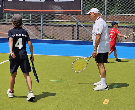 Älterer Trainer mit Tennisschläger und Ball neben Kindern auf dem Schwörsporttag