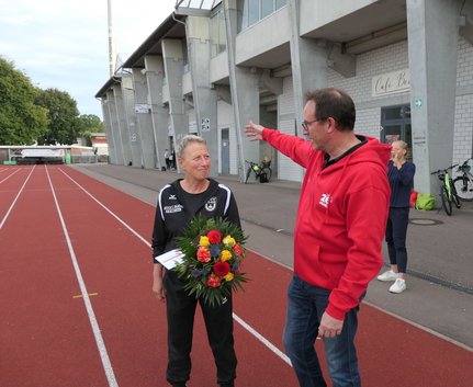 Frau mit Blumenstrauß und Umschlag auf Stadionlaufbahn neben gestikulierendem Mann