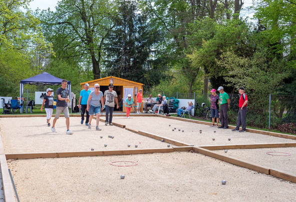 Mehrere Personen spielen Boule auf zwei Kiesbahnen vor einer Holzhütte
