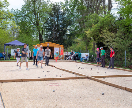 Mehrere Personen spielen Boule auf zwei Kiesbahnen vor einer Holzhütte