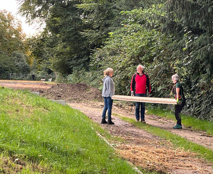 Drei Personen tragen ein langes Holzbrett auf Waldweg