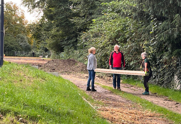 Drei Personen tragen ein langes Holzbrett auf Waldweg