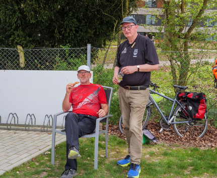 Zwei Männer neben Fahrrad, einer sitzt in rotem Shirt isst