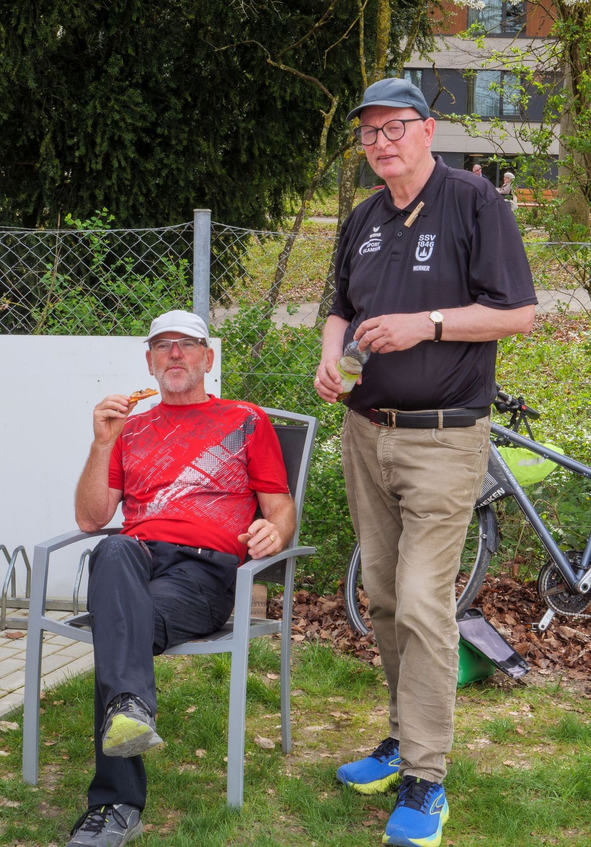 Zwei Männer neben Fahrrad, einer sitzt in rotem Shirt isst