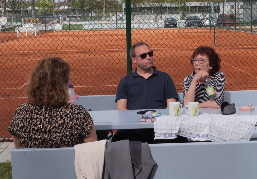 Drei Personen sitzen an Tisch vor rotem Tennisplatz