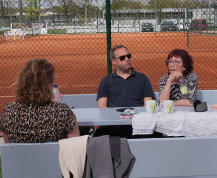 Drei Personen sitzen an Tisch vor rotem Tennisplatz