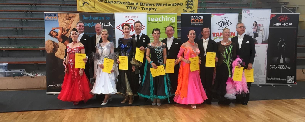 Tanzpaare in Abendkleidung mit Urkunden vor TBW-Trophy Banner