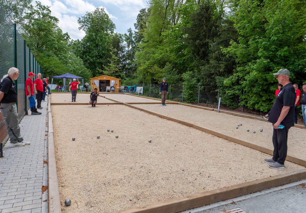 Menschen spielen Boule auf zwei Schotterbahnen neben Bäumen