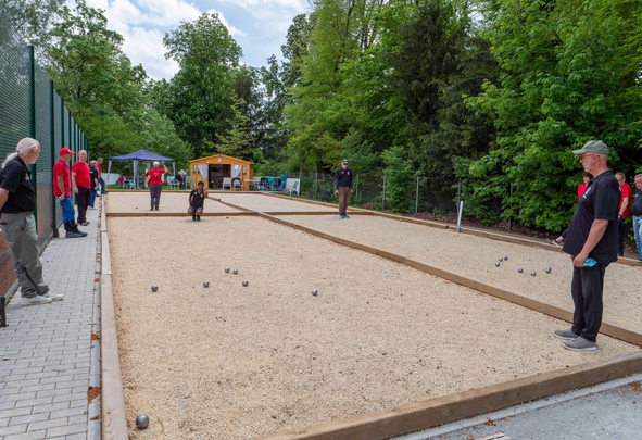 Menschen spielen Boule auf zwei Schotterbahnen neben Bäumen
