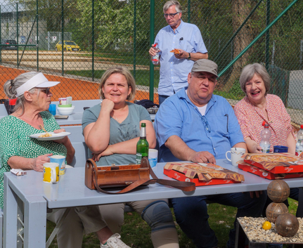 Fünf Erwachsene sitzen am Picknicktisch neben einem Tennisplatz mit Pizza