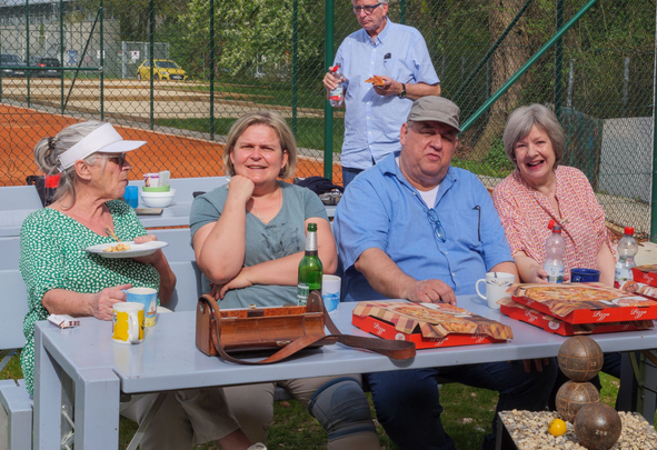 Fünf Erwachsene sitzen am Picknicktisch neben einem Tennisplatz mit Pizza