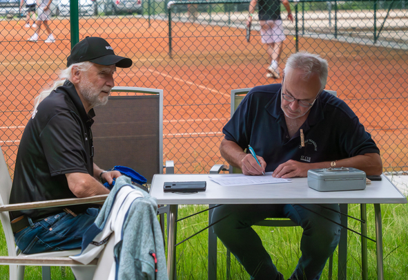 Zwei Männer sitzen am Tisch neben Tennisplatz, einer schreibt