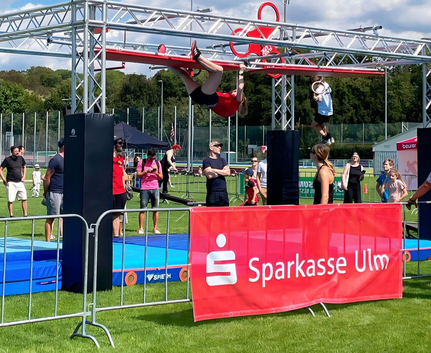 Chiara aus dem Team Sparkasse Ulm am letzten Hinderniss des Ninja Warrior Parcours Teilnehmende am Ninja-Warrior-Parcours rotes Banner Sparkasse Ulm im Vordergrund