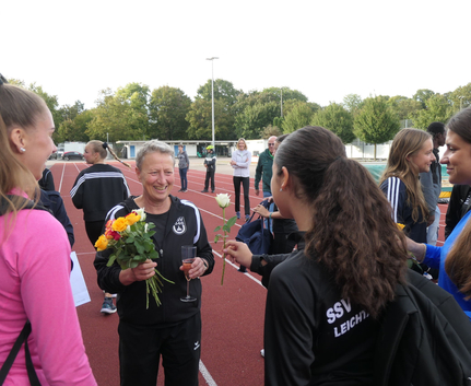 Brigitte lächelnd mit Blumen und Sektglas umgeben von U14 und U16 Athletinnen