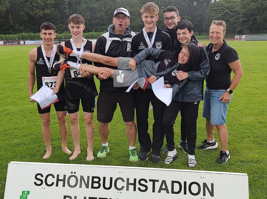 Hitze und Regen getrotzt: Die Ulmer Neunkampf-Riege U16-Athleten und Trainer tragen Teamkollegen vor Schild SCHÖNBUCHSTADION
