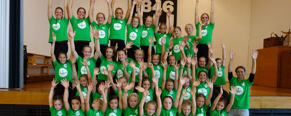 Lachende Mädchen in grünen T-Shirts mit erhobenen Händen beim Sommercamp