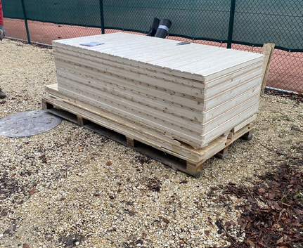 Gestapelte Holzplatten auf Palette neben grünem Tennisplatzzaun