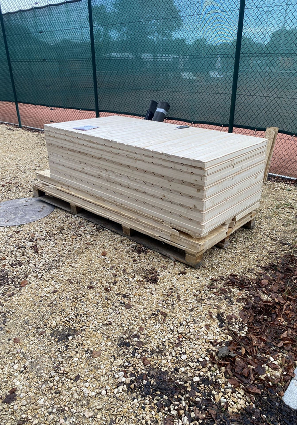 Gestapelte Holzplatten auf Palette neben grünem Tennisplatzzaun