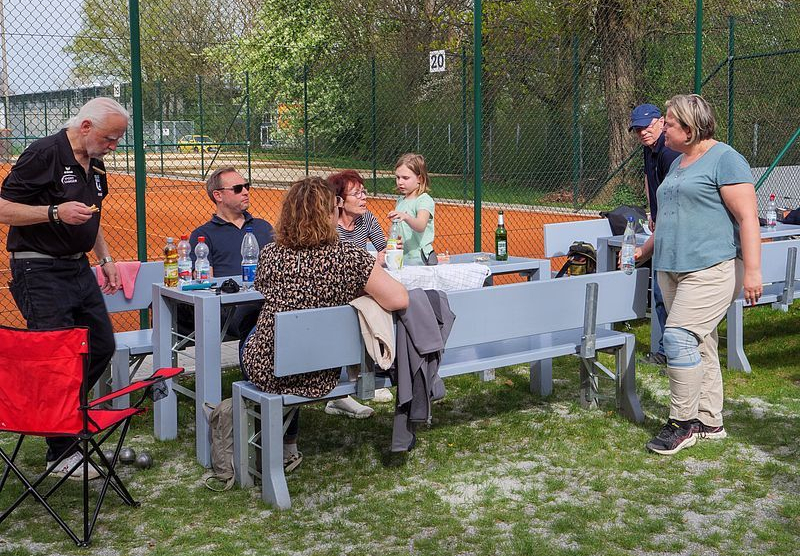 Mehrere Erwachsene und ein Mädchen an Tisch mit Bänken neben Tennisplatz