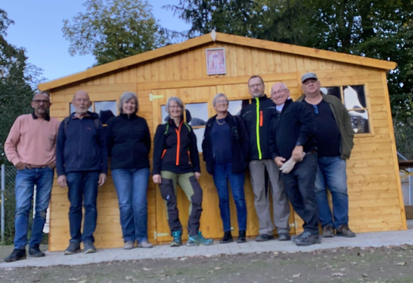 Acht Personen stehen vor einem hölzernen Gartenhaus im Freien
