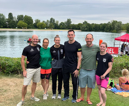 Sechs Personen in Sportkleidung am Seeufer mit Bojen und Zelt im Hintergrund