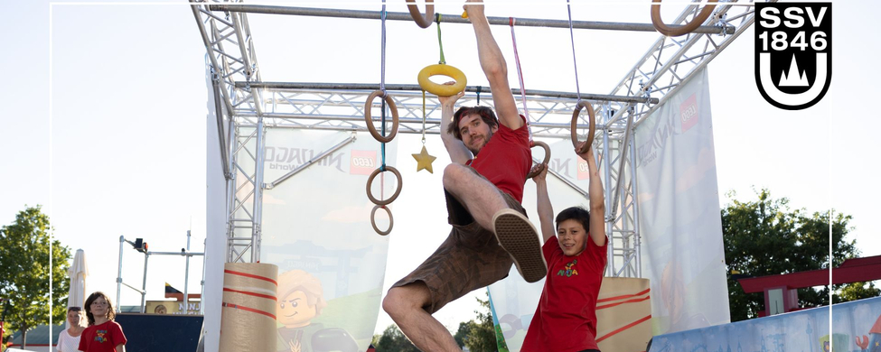 Erwachsener und Junge hängen an Ringen am Ninja Warrior Parcours auf Sporttag