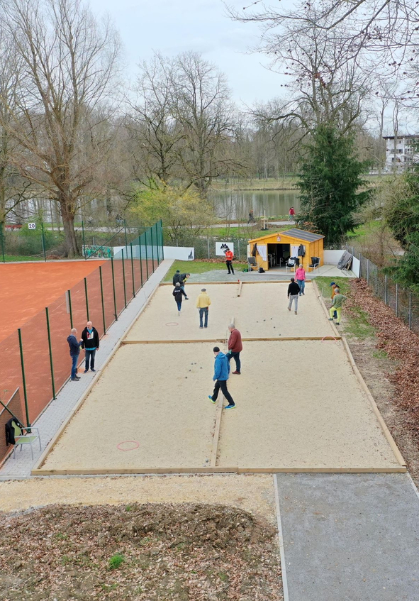 Mehrere Personen spielen Boule auf zwei Sandbahnen neben Tennisplatz