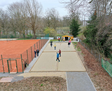 Mehrere Personen spielen Boule auf zwei Sandbahnen neben Tennisplatz