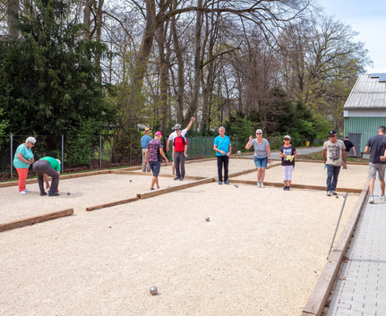 Mehrere Erwachsene spielen Boule auf Kiesbahnen unter Bäumen