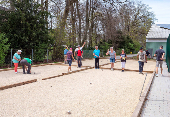 Mehrere Erwachsene spielen Boule auf Kiesbahnen unter Bäumen