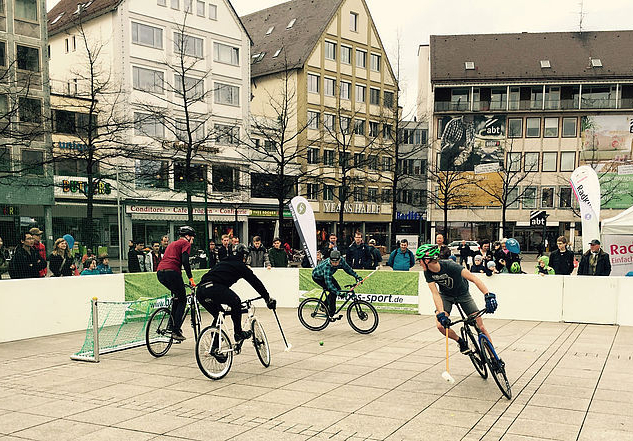 Fahrradballspieler mit Schlägern spielen auf gepflastertem Marktplatz vor Zuschauern