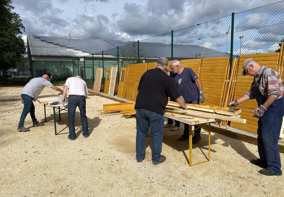 Fünf Männer arbeiten an Holzteilen auf Tischen im Freien