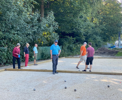 Sechs Männer spielen Boule auf gekiestem Platz vor Bäumen