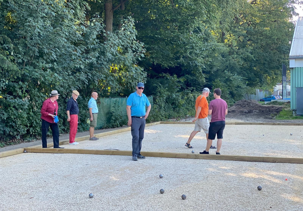Sechs Männer spielen Boule auf gekiestem Platz vor Bäumen