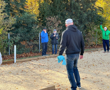 Gruppe auf Kiesplatz spielt Boule, eine Person hebt den Arm