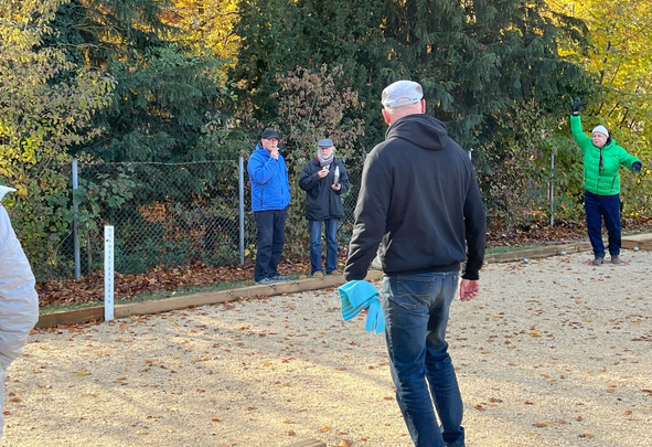 Gruppe auf Kiesplatz spielt Boule, eine Person hebt den Arm