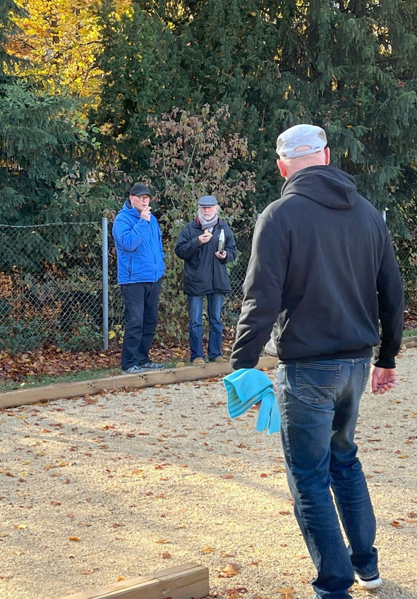 Gruppe auf Kiesplatz spielt Boule, eine Person hebt den Arm