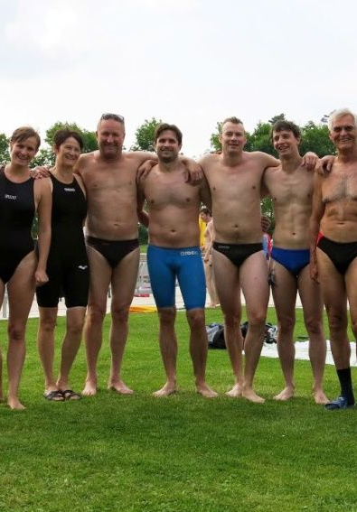 Elf Erwachsene in Badebekleidung stehen Arm in Arm auf Rasen neben Schwimmbad