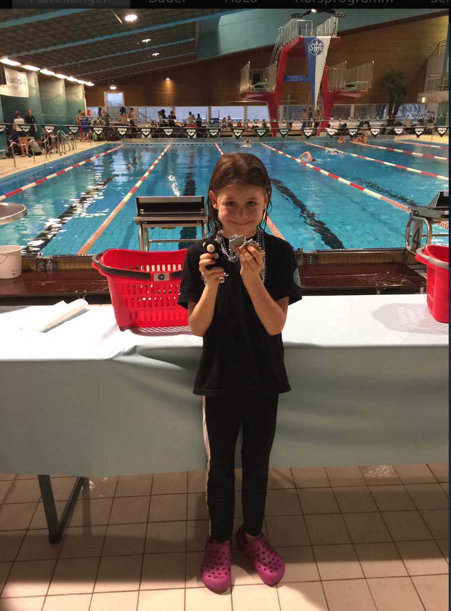 Kind vor Schwimmbecken hält Medaillen und kleine Trophäe