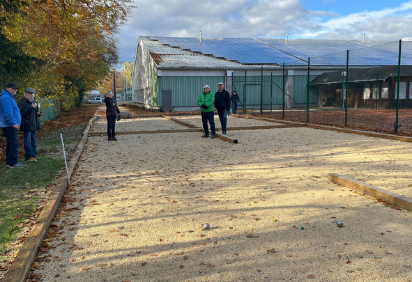 Mehrere Personen spielen Boule auf Kiesbahn vor Sporthalle