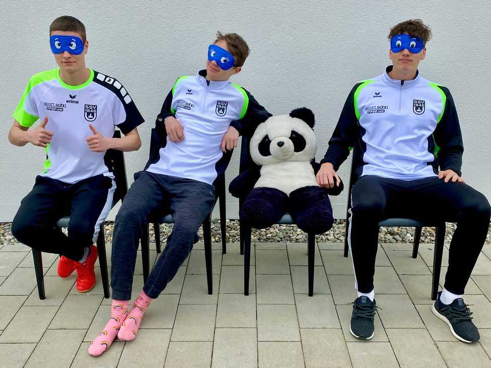 Drei Personen mit blauen Augenmasken sitzen neben großem Stoffpanda