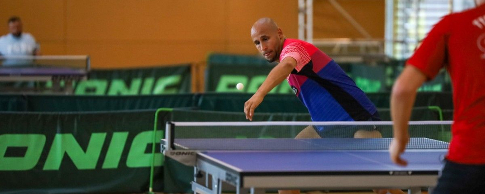 Tischtennisspieler in blauem Trikot greift Ball über den Tisch