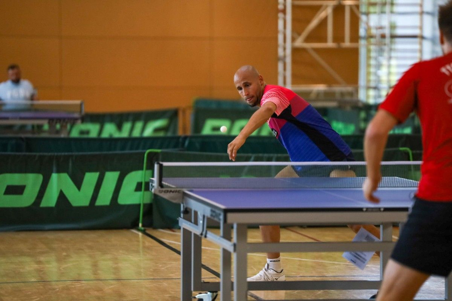 Tischtennisspieler in blauem Trikot greift Ball über den Tisch