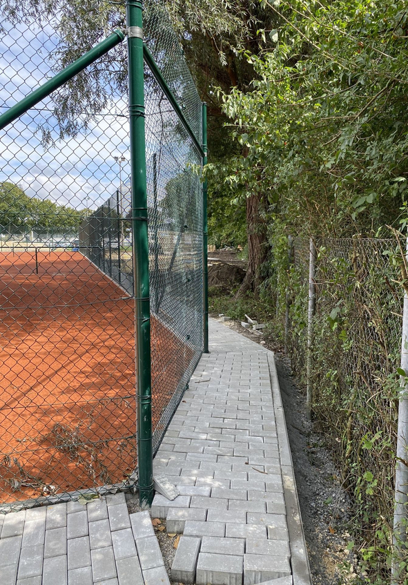 Pflasterweg neben grünem Maschendrahtzaun und rotem Tennisplatz