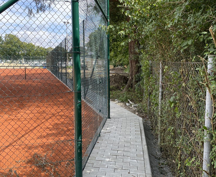 Pflasterweg neben grünem Maschendrahtzaun und rotem Tennisplatz
