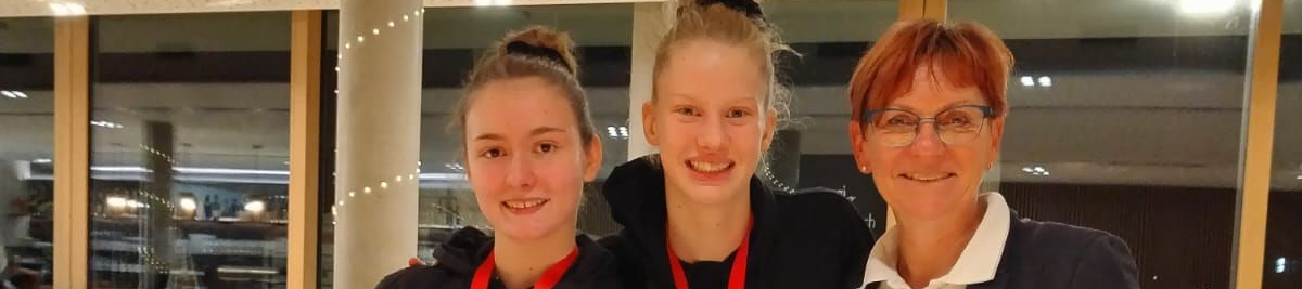 Zwei Gymnastinnen mit Medaillen und Trainerin in Innenraum