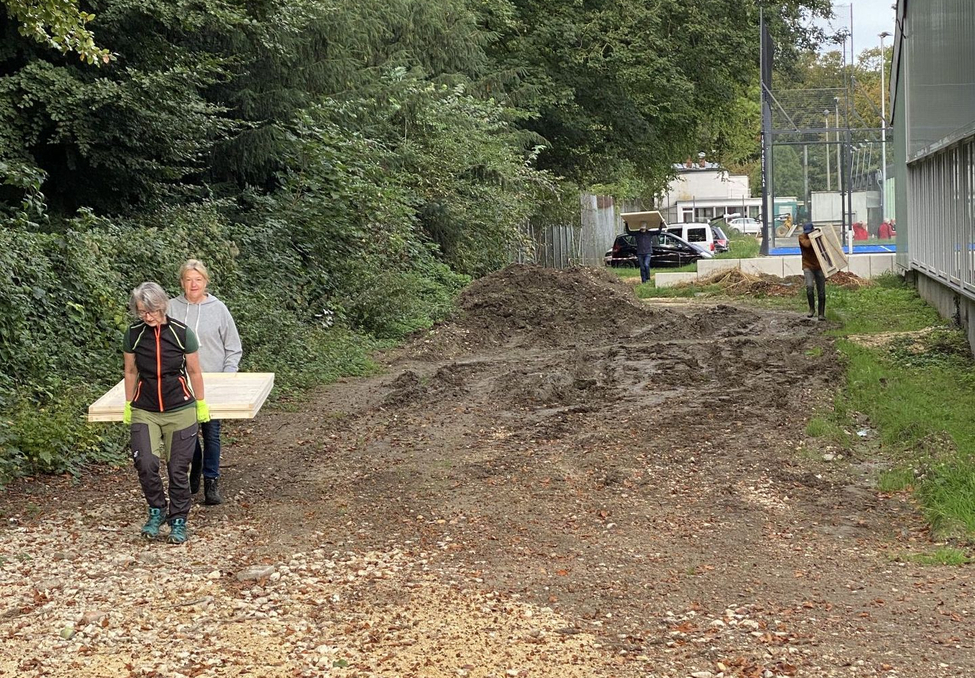 Zwei Personen tragen Holzbrett links, Person trägt Brett hinten auf Weg