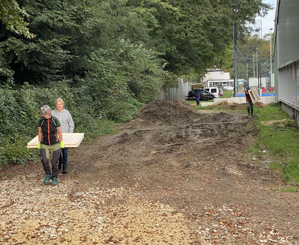 Zwei Personen tragen Holzbrett links, Person trägt Brett hinten auf Weg