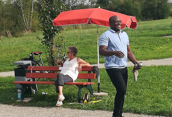 Lächelnder Mann in blauem Poloshirt neben Bank unter rotem Sonnenschirm